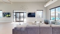 Located at 909 E Main St in the heart of Richmond, Virginia, this single-floor amenity space combines contemporary design with thoughtful functionality. The open lounge area serves as the centerpiece of the space, featuring soaring ceilings, recessed lighting, and an oversized sectional sofa arranged around a pair of round coffee tables. Large floor-to-ceiling windows and glass doors flood the room with natural light while offering sweeping views of the surrounding downtown cityscape. A wall-mounted flat-screen TV and decorative wall art add to the room's inviting character. Adjacent to the lounge, a dedicated dining area provides a substantial wooden table with seating for multiple guests, a wall-mounted display screen, and direct access to an outdoor terrace through full-height glass doors, perfect for seamless indoor-outdoor gatherings. The kitchen is a standout feature, finished in soft aqua tones with dark cabinetry, expansive white quartz countertops, open wood shelving, and stainless steel appliances including a double oven and French door refrigerator. A large central island with bar seating adds to the kitchen's practicality. Throughout, light-toned flooring ties the spaces together, creating a bright, cohesive, and modern urban retreat.
