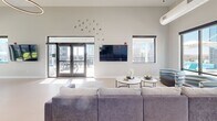 Located at 909 E Main St in the heart of Richmond, Virginia, this single-floor amenity space combines contemporary design with thoughtful functionality. The open lounge area serves as the centerpiece of the space, featuring soaring ceilings, recessed lighting, and an oversized sectional sofa arranged around a pair of round coffee tables. Large floor-to-ceiling windows and glass doors flood the room with natural light while offering sweeping views of the surrounding downtown cityscape. A wall-mounted flat-screen TV and decorative wall art add to the room's inviting character. Adjacent to the lounge, a dedicated dining area provides a substantial wooden table with seating for multiple guests, a wall-mounted display screen, and direct access to an outdoor terrace through full-height glass doors, perfect for seamless indoor-outdoor gatherings. The kitchen is a standout feature, finished in soft aqua tones with dark cabinetry, expansive white quartz countertops, open wood shelving, and stainless steel appliances including a double oven and French door refrigerator. A large central island with bar seating adds to the kitchen's practicality. Throughout, light-toned flooring ties the spaces together, creating a bright, cohesive, and modern urban retreat.