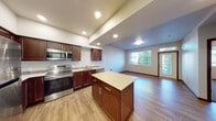 This single-floor unit offers a thoughtfully designed open-concept layout that seamlessly connects the kitchen and living area. The kitchen is a standout feature, showcasing rich dark wood cabinetry, light-toned countertops, and a full suite of stainless steel appliances including a range with over-the-range microwave, dishwasher, and refrigerator. A central kitchen island provides additional prep and storage space. Recessed lighting illuminates the kitchen, which transitions naturally into the open living area, where large windows and a glass entry door invite abundant natural light and outdoor views. Wood-look flooring runs throughout the main living spaces, lending warmth and continuity to the design. The bedroom features plush carpeting, a built-in shelf, and a spacious closet, offering a comfortable and functional retreat. The bathroom is well-appointed with a wide vanity, rich wood cabinetry, a large mirror, tile backsplash accents, and a tub-shower combination. A dedicated in-unit laundry closet houses a full-size stacked washer and dryer, adding everyday convenience. A separate utility or flex room with hardwood-style flooring and utility hookups rounds out this practical and comfortable living space.
