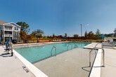 Located at 191 Commerce Dr in Villa Rica, Georgia, this outdoor amenity space serves as a central gathering hub for a modern multi-story residential community. The centerpiece is a generously sized rectangular swimming pool with crystal-clear water, accessible via stainless steel entry rails and surrounded by a wide, textured concrete deck designed for both safety and comfort. The pool area is well-equipped with rows of lounge chairs and chaise lounges positioned along multiple sides of the deck, providing ample space for sunbathing and relaxation. A shaded seating area with a blue umbrella offers a retreat from the sun, while string lights overhead add a welcoming ambiance during evening hours. A substantial natural wood bench serves as a bold architectural accent near the pool perimeter, complementing the contemporary design aesthetic. The entire area is enclosed by a black iron fence with a gated entry, ensuring safety and privacy. Surrounding landscaping includes ornamental grasses and young trees that add greenery to the open-air setting. The backdrop features the multi-story residential buildings with blue-gray siding, pitched rooflines, and private balconies. Open skies and views of the surrounding landscape enhance the spacious, resort-like feel of this well-appointed outdoor space.