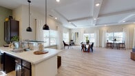 This single-floor community clubhouse is thoughtfully designed to offer a welcoming and functional gathering space. Light wood-look flooring runs throughout, complementing the warm, neutral color palette that lends the space a clean and contemporary feel.

The expansive lounge area is anchored by a coffered ceiling with recessed lighting, creating an elevated ambiance. A shiplap-accented feature wall hosts a wall-mounted television, while plush seating — including a leather sofa and teal accent chairs arranged around a coffee table — provides a comfortable setting for relaxation or socializing. Bar-height seating along the windows offers additional gathering spots bathed in natural light.

The open-plan layout flows seamlessly into a generous dining area, where a large wooden dining table with Windsor-style chairs sits beneath abundant natural light from multiple oversized windows dressed in sheer white curtains. A secondary seating nook with a round table and industrial-style chairs provides a more intimate setting for smaller groups. Original artwork adorns the walls, adding a curated touch throughout.

The kitchen is well-appointed with dark espresso cabinetry, white countertops, a central island with bar stool seating, pendant lighting, and full appliances — ideal for catered events or community gatherings. A connecting corridor links the various zones of the space efficiently, reinforcing its practical, community-oriented design.
