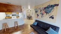 This single-floor residence offers a practical and inviting open-concept layout that seamlessly connects the living, dining, and kitchen areas. The living room features warm hardwood flooring and is anchored by a tufted black leather futon, complemented by a striking large-scale world map wall art piece that serves as a bold focal point. A ceiling fan overhead adds both function and casual charm to the space.

The kitchen is equipped with essential appliances including a white refrigerator and a range with overhead microwave. Warm wood-toned cabinetry lines the walls, and a raised breakfast bar with a wooden barstool provides a convenient spot for casual dining or meal prep. The kitchen transitions smoothly into the rest of the living area via a half-wall counter.

The dining area, situated just beyond the kitchen, features light tile flooring and hosts a compact dining table with chairs, making it ideal for everyday meals. A framed piece of wall art and a small decorative floral arrangement add a personal touch to the space.

Throughout the home, the combination of hardwood and tile flooring, neutral walls, and ceiling lighting create a clean, functional, and comfortable environment suited for everyday living.