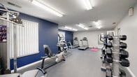 This fitness center is a thoughtfully designed, single-floor facility built to accommodate a wide range of workout needs. The space is defined by bold navy blue accent walls contrasted against light gray upper walls and a white ceiling, creating an energetic yet focused atmosphere. Durable rubber flooring runs throughout, providing both safety and comfort underfoot.

The cardio zone is well-stocked with multiple Matrix treadmills, an elliptical cross-trainer, and a recumbent exercise bike, offering users a variety of options for aerobic exercise. A dedicated stretching and floor exercise area features a yoga mat, providing space for warm-ups and cooldowns. Strength training is supported by a multi-station cable and weight machine, along with additional resistance equipment positioned along the perimeter.

Natural light filters into the room through several windows fitted with vertical blinds, supplemented by bright fluorescent overhead lighting that ensures the space remains well-lit at all times. A mirror is mounted on one wall, aiding in form correction during workouts. French doors with frosted glass panels connect to an adjacent area, adding an architectural touch while maintaining privacy. The overall layout is open and functional, allowing users to move freely between equipment stations.