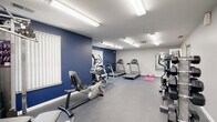 This fitness center is a thoughtfully designed, single-floor facility built to accommodate a wide range of workout needs. The space is defined by bold navy blue accent walls contrasted against light gray upper walls and a white ceiling, creating an energetic yet focused atmosphere. Durable rubber flooring runs throughout, providing both safety and comfort underfoot.

The cardio zone is well-stocked with multiple Matrix treadmills, an elliptical cross-trainer, and a recumbent exercise bike, offering users a variety of options for aerobic exercise. A dedicated stretching and floor exercise area features a yoga mat, providing space for warm-ups and cooldowns. Strength training is supported by a multi-station cable and weight machine, along with additional resistance equipment positioned along the perimeter.

Natural light filters into the room through several windows fitted with vertical blinds, supplemented by bright fluorescent overhead lighting that ensures the space remains well-lit at all times. A mirror is mounted on one wall, aiding in form correction during workouts. French doors with frosted glass panels connect to an adjacent area, adding an architectural touch while maintaining privacy. The overall layout is open and functional, allowing users to move freely between equipment stations.