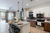 This well-appointed single-floor residence showcases a thoughtfully designed open-concept layout with modern finishes throughout. The kitchen serves as a central hub, featuring a large granite-topped island with bar seating, crisp white upper cabinetry, warm wood-toned lower cabinets, and a striking dark subway tile backsplash. Stainless steel appliances — including a side-by-side refrigerator, range, microwave, and dishwasher — complete the space. Light wood-style flooring flows throughout, tying each area together cohesively.

The living area offers a comfortable, light-filled retreat with large windows dressed in layered drapes, ceiling fans, and ample room for relaxing or entertaining. Multiple bedrooms provide versatile accommodations, each with ceiling fans, natural light from generous windows, and the same warm wood flooring. The primary bedroom benefits from its own well-designed en suite bathroom, featuring a granite vanity with wood cabinetry, a large framed mirror, and a glass-enclosed walk-in shower with white subway tile. A secondary bathroom mirrors the contemporary aesthetic with a granite vanity and a tub-shower combination with subway tile surround. The overall design balances clean, neutral tones with warm wood accents for a modern yet inviting ambiance.