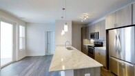 This single-floor residence at Overture showcases a thoughtfully designed open-concept layout that seamlessly connects the kitchen and living areas. The kitchen serves as the centerpiece, anchored by a generous island topped with light-toned granite countertops that feature subtle veining and a polished finish. The island includes an undermount sink with a modern arc faucet and is illuminated by stylish pendant lights with warm brass accents, adding a refined touch to the space.

The kitchen cabinetry features a light wood-grain finish in a contemporary flat-panel style, complemented by a subway tile backsplash and stainless steel appliances, including a range and over-the-range microwave. Track lighting above the cooking area provides additional task illumination.

The adjoining living area is bright and airy, with multiple windows allowing ample natural light to fill the room. A ceiling fan enhances comfort in the living space. Consistent hardwood-style flooring flows throughout, unifying the open layout. Neutral gray walls and white ceilings contribute to the clean, modern aesthetic, creating a welcoming and functional living environment.