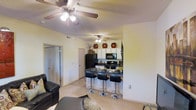 This single-floor residence is designed with an open-concept layout that connects the living area and kitchen into one cohesive, functional space. The living area is finished with light-toned carpet and features a ceiling fan with integrated lighting, contributing to a comfortable and well-ventilated atmosphere. A leather sofa and area rug anchor the lounge space, which flows naturally toward the kitchen without visual obstruction.

The kitchen is equipped with black appliances, including a refrigerator, range, and microwave, set against white cabinetry that extends to the upper walls. A dark-surfaced breakfast bar separates the kitchen from the living space and is accompanied by sleek, chrome-accented bar stools, offering casual seating for dining or entertaining. Upper cabinet space is maximized with decorative storage above the cabinetry. A second ceiling fan is positioned over the kitchen area, ensuring consistent air circulation throughout the open floor plan.

A hallway visible near the entrance suggests access to additional rooms, and the overall layout reflects a practical, well-organized single-level design suited for everyday living.