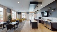 This well-appointed community amenity space combines a fully equipped kitchen and lounge area into a single, open-concept layout that encourages both social interaction and focused work. The kitchen is anchored by a large central island with bar seating and overhead pendant lighting, complemented by rich dark cabinetry, a tiled backsplash, white countertops, and stainless steel appliances including a beverage station and under-counter refrigerator. A wall-mounted flat-screen television and decorative open shelving add both function and character to the kitchen wall. Adjacent to the kitchen, the lounge area features light hardwood-style flooring that transitions seamlessly into patterned area rugs. Seating arrangements include a navy blue sofa, upholstered yellow accent chairs with geometric patterning, and a variety of mixed dining and café-style tables with chairs, offering flexible options for gatherings of all sizes. A dedicated nook near large windows provides a quieter setting with additional yellow armchairs, ideal for casual meetings or individual use. Throughout the space, floor-to-ceiling windows dressed with neutral drapes allow generous natural light to pour in while offering views of the surrounding area. Recessed lighting and statement pendant fixtures complete the polished, contemporary aesthetic. A large decorative wall-mounted Scrabble board adds a playful, communal touch to the overall design.