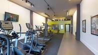 This contemporary fitness center is thoughtfully designed to provide a complete workout environment within a single, elongated space. The room is outfitted with rubber flooring throughout, offering both durability and comfort underfoot during exercise. Track lighting runs along the ceiling, casting focused, even illumination across the entire floor, while recessed lights further enhance the bright and energizing atmosphere.

The space is equipped with a range of cardio machines, including ellipticals and other aerobic equipment, positioned along one side of the room to allow for a natural flow of movement. Toward the rear, a multi-station strength training machine provides a centralized hub for resistance-based workouts. A striking yellow accent wall anchors the far end of the space, adding a bold, motivating pop of color that contrasts with the neutral grey and white tones dominating the rest of the interior.

Full-length mirrors are mounted along the walls, visually expanding the space and allowing users to monitor their form during exercise. The layout is open and uncluttered, supporting ease of movement between stations. A private room or storage area is accessible through a doorway at the far end, adding functional versatility. Overall, the space strikes a balance between energetic design and practical functionality.