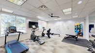 Located at 7880 Alpha Rd in Dallas, TX, this fitness center offers a well-appointed exercise space designed to support a full range of workout routines. The single-floor layout is efficiently organized, comprising a main exercise room and a connecting hallway accessed through glass-panel double doors.

The exercise room is bright and clean in its design, featuring a drop-panel ceiling equipped with recessed and panel lighting, as well as a ceiling fan for added airflow and comfort. Neutral-toned, multi-shade patterned tile flooring runs throughout, lending a modern, professional aesthetic. Natural light enters through multiple windows, offering views of the surrounding outdoor greenery and Dallas skyline.

The room is furnished with a variety of cardio equipment, including treadmills, an elliptical, a stationary bike, and a rowing machine. A weight bench and a rack of dumbbells provide strength-training options, while a padded floor mat creates a dedicated area for stretching or floor exercises. A large wall-mounted flat-screen TV is positioned for easy viewing during workouts.

A striking geometric mural of a running athlete spans one wall, serving as a bold visual focal point. Full-length mirrors on an adjacent wall amplify the sense of space. The hallway area features light hardwood-style flooring and an additional wall-mounted TV, completing this functional and visually engaging fitness facility.