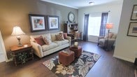 This single-floor residence offers a cohesive and well-appointed interior defined by rich hardwood flooring, crisp crown molding, and a warm, earthy color palette. The living area serves as the heart of the space, showcasing neutral walls accented by a deep taupe feature wall that adds depth and sophistication. A generously sized sofa anchors the room, complemented by rust-toned ottomans and a boldly patterned area rug with floral motifs in earthy browns and creams. Decorative framed artwork adorns the walls, adding a curated, homey touch alongside a classic wall clock. Natural light filters in through large windows dressed with dark curtain panels, creating a balanced interplay of light and shadow. A compact built-in desk area with shelving provides a functional workspace seamlessly integrated into the living space, making it ideal for those who work from home. The overall ambiance is warm, grounded, and inviting, with carefully chosen furnishings and décor that create a comfortable yet stylish environment. The single-level layout ensures easy accessibility and a fluid sense of continuity throughout the home.