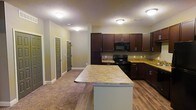 This single-floor space offers an open-concept layout connecting the kitchen and living areas seamlessly. The kitchen is a standout feature, showcasing rich dark espresso cabinetry that extends to the upper walls, providing generous storage throughout. Stone-look countertops in warm golden and brown tones complement the cabinetry, running along the perimeter and continuing onto a central kitchen island, which adds additional prep space and serves as a natural divider between the kitchen and the adjoining living area. Black appliances, including a range with an over-the-range microwave, complete the kitchen's cohesive, contemporary look. The flooring transitions from hardwood-style planks in the kitchen to plush carpet in the living area, adding texture and warmth. Flush-mounted ceiling light fixtures illuminate the space evenly throughout. The textured ceiling adds subtle dimension overhead. Multiple interior doors are visible along the back wall, suggesting access to additional rooms or closets. Neutral wall tones in a soft sage-green hue provide a calm, understated backdrop that ties the kitchen and living spaces together cohesively.