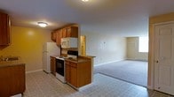 This single-floor space offers an open and functional layout that seamlessly connects the kitchen and living area. The kitchen is equipped with warm honey-toned wood cabinetry, both upper and lower, providing ample storage. A peninsula counter with a dark laminate surface serves as a natural divider between the kitchen and the adjoining living space, offering additional prep area or casual seating potential. Essential appliances are included, among them a white refrigerator, an electric range with oven, and an over-the-range microwave. The kitchen floor features a light-toned tile with a subtle pattern, keeping the space bright and easy to maintain. Transitioning into the living area, the flooring shifts to wall-to-wall carpeting in a neutral tone, creating a comfortable and cohesive feel underfoot. The living space is generously proportioned, with smooth walls painted in a soft, warm neutral palette that enhances the sense of openness. Recessed ceiling lighting in the kitchen and a flush-mount fixture provide functional illumination throughout. A hallway visible from the kitchen suggests access to additional rooms, completing this practical and well-organized single-floor layout.