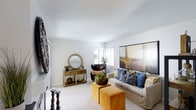 Located in San Diego on Avenida Alcochofa, this single-story home offers a well-appointed living space designed with both comfort and style in mind. The living room serves as the focal point of the home, showcasing smooth white ceilings that enhance the sense of openness and light throughout the space. Soft, neutral-toned carpet underfoot adds warmth and complements the overall color palette.

Natural light filters in through windows dressed with slatted blinds, illuminating the room's carefully curated décor. The seating area features a classic cream sofa accented with a rich mix of blue patterned and gold throw pillows, paired with a sleek black accent chair. A pair of plush mustard-yellow ottomans adds a bold pop of color and functional versatility to the arrangement.

Decorative touches elevate the space further, including a large sunburst mirror above a wooden console table styled with greenery and decorative objects. A substantial piece of abstract wall art anchors one wall, while a bold circular wall sculpture adds artistic depth to another. The overall ambiance is polished yet approachable, blending contemporary and eclectic design elements seamlessly.