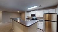 Located at Cameron Woods Drive in Apex, NC, this single-floor residence offers a well-designed layout with a clean, modern aesthetic throughout. The kitchen is a functional highlight, featuring white shaker-style cabinetry, dark granite-look countertops, a classic subway tile backsplash, and a full suite of stainless steel appliances including a refrigerator, electric range, and over-the-range microwave. A peninsula with bar-top seating connects the kitchen to the living area, creating an open and social flow. Light wood-look flooring runs through the kitchen, providing a warm contrast to the crisp cabinetry. The spacious living area is finished with plush neutral carpeting and is brightened by a ceiling fan light fixture and access to a door leading to the outdoors, allowing natural light to filter in through large windows. The bedrooms are generously proportioned, each with soft carpeting, neutral wall tones, and windows overlooking lush greenery, creating a calm and restful atmosphere. The bathrooms are neatly appointed with white tile tub-shower combinations, dark countertop vanities with white cabinetry, and wood-look flooring. Overall, the space presents a cohesive, move-in-ready interior with consistent finishes and a practical, comfortable design.