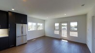 This contemporary single-floor space offers a clean, modern aesthetic defined by crisp white walls, smooth ceilings with recessed lighting, and rich dark hardwood flooring throughout. The open-concept layout creates a seamless flow between the living and kitchen areas, lending a spacious and airy feel to the interior. Large horizontal windows line the walls, inviting generous amounts of natural light and offering views of the surrounding greenery. A set of elegant French doors provides direct access to an outdoor area, further enhancing the connection between the interior and the outside environment. The kitchen area is equipped with sleek dark upper cabinetry and a stainless steel refrigerator, offering a striking contrast against the bright white walls. Recessed ceiling lights complement the abundant natural light, ensuring the space remains well-illuminated at all times. The overall ambiance is open, clean, and contemporary, making the space feel welcoming and functional. The minimalist design approach, combined with quality finishes and thoughtful architectural details, creates a polished and versatile environment suitable for a variety of uses.