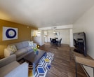 This single-floor residence offers a thoughtfully designed layout that balances comfort and functionality across its key living spaces. The open-plan living and dining area serves as the heart of the home, featuring durable wood-style flooring, a warm gold accent wall, and a sliding glass door that invites natural light and provides access to an outdoor patio with surrounding greenery. The adjoining dining area is illuminated by a pendant light fixture, creating a welcoming atmosphere for meals and gatherings. The galley-style kitchen is efficiently arranged with white cabinetry, light countertops, a gas range, dishwasher, refrigerator, and a built-in microwave. Track lighting brightens the workspace, and a dedicated walk-in pantry offers generous storage. Two well-proportioned bedrooms feature plush carpeting, natural window light, and ample closet space. The primary bedroom includes a ceiling fan and access to a walk-in closet. Both bathrooms are finished with light countertops, white cabinetry, and wood-style flooring — one featuring a full tub-shower combination with a well-lit vanity mirror. Completing the unit is a dedicated laundry room equipped with a full-size washer and dryer beneath generous upper cabinetry storage.