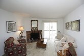 This single-floor residence offers a well-organized layout designed for comfortable everyday living. The living room serves as the central gathering space, highlighted by a classic brick fireplace and a sliding glass door that fills the room with natural light and provides access to an outdoor area. Soft carpeting and neutral walls create a warm, inviting ambiance throughout.

The kitchen is efficiently arranged with rich wood-toned cabinetry offering generous storage above and below the countertops. It is equipped with a range, dishwasher, and a deep sink, making it a practical space for meal preparation. Light tile flooring completes the functional design.

Two bedrooms provide comfortable retreats, each featuring plush carpeting, natural light from large windows, and closet storage. The primary bedroom is notably spacious, with room for a full dresser and nightstands. The secondary bedroom is equally well-lit and includes an open closet with shelving.

The bathroom features an extended vanity with dual mirror configurations, dark wood cabinetry below a white countertop, and tiled flooring. A separate tub and shower area adds practicality. Throughout the residence, textured ceilings and a consistent neutral color palette lend a cohesive, understated character.