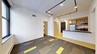 This contemporary single-floor space boasts a clean, open-concept design that seamlessly connects the living area to the kitchen. The living area is finished with distinctive patterned carpet tiles featuring bold yellow accents, adding a modern, design-forward touch to the floor. Crisp white walls and high ceilings amplify the sense of openness, while industrial-style black track lighting runs across the ceiling, providing flexible and stylish illumination throughout.

The kitchen is positioned along one side of the open space and features warm wood-toned cabinetry, a dark countertop island with an integrated sink, and stainless steel appliances including a refrigerator. A pendant light adds an elegant focal point above the kitchen area, complementing the overall aesthetic.

A dedicated laundry area is accessible through a doorway off the main living space, housing a stacked washer and dryer unit for added convenience. Additional doors lead to a closet and what appears to be a bathroom or private room, offering functional separation within the layout. The combination of neutral tones, modern lighting, and thoughtful finishes creates a clean, urban ambiance suited for contemporary living.