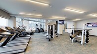 This fitness center offers a comprehensive workout environment designed to accommodate a variety of exercise routines. The space is organized across a single floor with an open, L-shaped layout that promotes ease of movement and clear sightlines throughout.

The cardio zone is prominently positioned along a large floor-to-ceiling window wall, flooding the area with abundant natural light and offering views of the surrounding urban streetscape. A row of treadmills, elliptical machines, and stair climbers lines this bright perimeter, creating an energizing atmosphere for endurance training.

Moving deeper into the space, a robust selection of strength and resistance training equipment is thoughtfully arranged, including cable machines, weight benches, and various selectorized units. A mounted flat-screen television provides entertainment and keeps members informed during workouts.

The interior walls feature exposed brick accents, adding a warm, industrial character that contrasts with the clean white ceilings fitted with long, linear LED lighting panels. The flooring throughout consists of durable, textured carpet tiles suited for gym use.

A dedicated seating and lounge area with a round table and chairs is situated in one corner of the facility, offering a space for rest or casual gatherings. The overall ambiance is bright, modern, and functional, making this fitness center a well-rounded amenity.