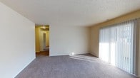 This single-floor residence presents a clean, functional layout with a warm and understated aesthetic throughout. The main living area is finished with wall-to-wall carpeting in a neutral gray tone, providing a soft and comfortable underfoot feel. Walls are painted in a light beige, contributing to a bright and welcoming atmosphere. A large vertical-blind window allows generous natural light to fill the space, casting a warm glow across the room.

The textured popcorn ceiling adds a classic residential character, while white baseboards provide a clean contrast against the carpeted floors. An open passageway connects the living area to an adjoining hallway, which leads further into the home. The hallway reveals warm yellow-toned walls, a ceiling fan fixture for added ventilation, and what appears to be a closet with bi-fold doors, suggesting convenient built-in storage solutions.

The overall layout is straightforward and practical, making efficient use of the available space. The neutral color palette and soft materials create a calm, livable environment suited for everyday comfort. Natural light and the open flow between rooms enhance the sense of spaciousness throughout this single-level home.