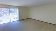This single-floor space offers a clean and functional layout with a welcoming, understated aesthetic. The main living area is finished with wall-to-wall carpeting in a neutral tone, providing a soft and comfortable underfoot feel throughout. Smooth, light-colored walls create a calm and airy atmosphere, while classic wood-toned baseboards add a subtle finishing detail to the room's perimeter.

A standout feature of the living area is the full-height sliding glass door fitted with vertical blinds, which allows abundant natural light to pour into the space. The door opens directly to an outdoor balcony, offering views of surrounding greenery and architectural elements beyond — ideal for enjoying fresh air and natural scenery.

The overall ambiance is bright and neutral, making the space highly adaptable for a variety of furnishing styles and personal tastes. The open layout of the main room provides generous flexibility for furniture arrangement, accommodating both lounging and functional living needs with ease. The combination of natural light, soft carpeting, and a connected outdoor space gives this residence a comfortable and livable character.