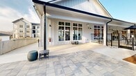 This impressive outdoor space is designed for ultimate leisure and entertainment. The centerpiece is a beautifully crafted stone grill station, featuring multiple built-in grills and ample counter space for food preparation. Overhead, a robust wooden pergola adds architectural interest and provides partial shade. The area is further enhanced by string lights, creating a warm ambiance for evening gatherings.

The expansive patio is laid with high-quality, large-format pavers in a neutral tone, providing a smooth and stylish surface. Comfortable seating options, including modern gray outdoor furniture, invite relaxation and socialization. The space is thoughtfully bordered by a combination of stone retaining walls and metal fencing, ensuring privacy while maintaining an open feel.

In the background, multi-story residential buildings are visible, suggesting this is a shared community space in an upscale apartment or condominium complex. The surrounding landscape includes mature trees, adding natural beauty and shade to the area. This meticulously designed outdoor retreat seamlessly blends functionality with aesthetic appeal, offering residents a sophisticated space for outdoor cooking, dining, and entertainment throughout the seasons.