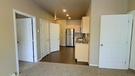 This single-story unit offers a practical and well-organized layout that transitions seamlessly from the carpeted living area into the kitchen and hallway beyond. The kitchen is equipped with stainless steel appliances, including a full-sized refrigerator and range, complemented by light wood cabinetry and a granite-style countertop with an integrated sink. Recessed lighting illuminates the kitchen and hallway, lending a clean, modern feel to the space. Dark hardwood-style flooring runs through the kitchen and corridor, providing a contrast to the carpeted living area and contributing to a cohesive aesthetic throughout. The hallway leads to multiple white-paneled doors, suggesting access to additional rooms or storage, all arranged in a tidy, functional corridor. Neutral beige wall tones and elevated ceilings enhance the sense of openness, creating a bright and airy atmosphere. The overall design prioritizes practicality while maintaining a clean, contemporary appearance suited for everyday living.