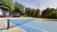 This property features outdoor tennis courts finished in a classic blue hard-court surface with crisp white boundary lines and a center net. The courts are enclosed by chain-link fencing and equipped with tall light poles, allowing for evening play. Mature trees surround the perimeter, providing a natural backdrop and partial shade, while an open sky above ensures excellent visibility during daytime use.

The adjacent interior living space offers a comfortable, functional layout. The main living area is carpeted and features a fireplace with a clean white surround, creating a focal point well-suited for relaxing after outdoor activity. A sliding glass door fitted with vertical blinds allows natural light to filter into a cozy seating nook, which connects the interior to views of the surrounding greenery outside.

The space also includes a hallway connecting additional rooms, with a textured ceiling, built-in closet storage, and wall-mounted thermostat controls. The entryway, finished with hardwood-style flooring and a dark front door, provides a clear transition between the interior and exterior. Overall, the space balances active outdoor amenities with a calm, understated interior atmosphere.
