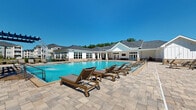 This single-level outdoor amenity complex is designed to deliver a resort-style experience, centered around a generously sized swimming pool with crystal-clear turquoise water. The pool is framed on multiple sides by a broad, stone-paver deck that provides ample space for lounging, with rows of cushioned chaise lounges and side tables arranged throughout. A water feature adds a dynamic focal point at one end of the pool, while a pergola structure at the far end offers shaded seating and additional gathering space.

The adjacent clubhouse building is finished in crisp white board-and-batten siding with a dark pitched roof and large multi-pane windows that allow natural light to flow both inside and out. An expansive covered outdoor patio extends from the clubhouse, featuring high ceilings equipped with ceiling fans, stone-tile flooring, and comfortable modular seating arranged around a wall-mounted display screen. French doors connect the covered patio seamlessly to the interior, blurring the line between indoor and outdoor living.

Brick-accented columns and auxiliary outbuildings complement the white exterior palette, adding warmth and architectural texture to the overall design. The surrounding landscape includes mature trees and palm trees, contributing to a lush, open-air atmosphere. Under abundant natural light and clear blue skies, this facility offers a polished and inviting environment suited for relaxation and social gatherings alike.