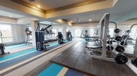 This fitness center is thoughtfully designed to accommodate a full range of workout needs across a generously proportioned, single-level space. The layout is organized into distinct functional zones, each outfitted with purpose-specific equipment and flooring. Striped carpet tiles in teal, beige, and gray tones define the cardio areas, while rubber flooring anchors the strength training and free weight zones, providing both durability and safety.

The cardio section houses multiple treadmills, upright and recumbent stationary bikes, ellipticals, and a rowing machine. Large windows throughout this area invite abundant natural light and offer views of the surrounding exterior, creating an open and energizing atmosphere. Ceiling fans are positioned throughout to promote airflow and comfort during workouts.

The strength training area features a cable machine, a weight rack with a comprehensive dumbbell selection, a barbell rack with weight plates, and adjustable benches. Full-length mirrors line the walls in this zone, enhancing spatial perception and allowing users to monitor their form. Recessed lighting supplements natural light throughout the facility, and a wall-mounted television is present for added convenience. The overall design is clean, modern, and functional.