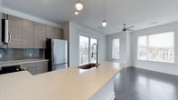 This single-floor space showcases a thoughtfully designed open-concept layout that seamlessly connects the kitchen and living areas. The kitchen is a standout feature, anchored by a generous island with a light-toned quartz countertop that provides ample workspace and a built-in undermount sink with a bronze gooseneck faucet. Overhead, stylish globe pendant lights hang above the island, adding a modern decorative touch. Cabinetry in a warm wood-grain finish lines the walls, complemented by stainless steel appliances, including a full-size refrigerator. Dark hardwood-style flooring runs throughout, providing a striking contrast to the light grey walls and bright countertops. The living area benefits from multiple windows fitted with blinds, allowing generous natural light to fill the space. A ceiling fan with a brushed nickel finish is centrally positioned in the living area, adding both function and a clean aesthetic. The overall ambiance is contemporary and airy, with a neutral color palette and modern fixtures that create a cohesive, polished feel throughout.