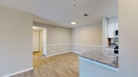 This single-floor residence features a thoughtfully designed open-concept layout with distinct living, dining, and kitchen zones that flow seamlessly together. Light-toned luxury vinyl plank flooring runs throughout the main living areas, complementing the neutral gray and beige wall palette and white baseboards accented by a decorative chair rail.

The living area is bright and open, with access to an exterior door and a textured ceiling overhead. It connects directly to the dining space, which is bordered by a peninsula-style kitchen counter featuring granite countertops. The kitchen is well-appointed with white cabinetry, granite countertops, a tile backsplash, and a full suite of stainless steel appliances including a refrigerator, dishwasher, over-the-range microwave, and electric range.

The bedroom offers plush wall-to-wall carpeting, a ceiling fan with light fixture, and a window with blinds that brings in natural light and outdoor views. The bathroom is finished with wood-look tile flooring, a white vanity topped with granite, and a full tub-shower combination. Overall, the space presents a clean, modern aesthetic with consistent finishes throughout.