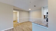 This single-floor residence features a thoughtfully designed open-concept layout with distinct living, dining, and kitchen zones that flow seamlessly together. Light-toned luxury vinyl plank flooring runs throughout the main living areas, complementing the neutral gray and beige wall palette and white baseboards accented by a decorative chair rail.

The living area is bright and open, with access to an exterior door and a textured ceiling overhead. It connects directly to the dining space, which is bordered by a peninsula-style kitchen counter featuring granite countertops. The kitchen is well-appointed with white cabinetry, granite countertops, a tile backsplash, and a full suite of stainless steel appliances including a refrigerator, dishwasher, over-the-range microwave, and electric range.

The bedroom offers plush wall-to-wall carpeting, a ceiling fan with light fixture, and a window with blinds that brings in natural light and outdoor views. The bathroom is finished with wood-look tile flooring, a white vanity topped with granite, and a full tub-shower combination. Overall, the space presents a clean, modern aesthetic with consistent finishes throughout.
