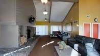 This single-floor space makes a strong architectural statement with its soaring vaulted ceiling accented by exposed wooden beams and a white-painted wood plank finish. A series of pendant light fixtures hang at varying heights along the ceiling's ridge, adding both functionality and visual interest to the room's vertical space.

The living area is anchored by a commanding floor-to-ceiling stone fireplace on one wall, constructed with rugged natural stonework and flanked by a stacked firewood storage nook, giving the space a rustic, lodge-like character. A wall-mounted television is positioned above the fireplace for convenience.

The walls are painted in a warm golden yellow, creating an inviting and energetic ambiance throughout. Large windows dressed with white vertical blinds allow generous natural light to fill the room. The space is furnished with dark leather sofas and chairs arranged around a central coffee table, offering a comfortable and cohesive seating arrangement. A framed artwork piece adds a decorative focal point on one wall. The carpeted flooring complements the warm tones of the overall interior, contributing to a cozy yet spacious atmosphere.