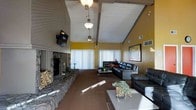 This single-floor space makes a strong architectural statement with its soaring vaulted ceiling accented by exposed wooden beams and a white-painted wood plank finish. A series of pendant light fixtures hang at varying heights along the ceiling's ridge, adding both functionality and visual interest to the room's vertical space.

The living area is anchored by a commanding floor-to-ceiling stone fireplace on one wall, constructed with rugged natural stonework and flanked by a stacked firewood storage nook, giving the space a rustic, lodge-like character. A wall-mounted television is positioned above the fireplace for convenience.

The walls are painted in a warm golden yellow, creating an inviting and energetic ambiance throughout. Large windows dressed with white vertical blinds allow generous natural light to fill the room. The space is furnished with dark leather sofas and chairs arranged around a central coffee table, offering a comfortable and cohesive seating arrangement. A framed artwork piece adds a decorative focal point on one wall. The carpeted flooring complements the warm tones of the overall interior, contributing to a cozy yet spacious atmosphere.