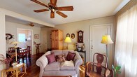 This single-floor, one-bedroom, one-bathroom residence offers a warm and inviting atmosphere with a distinctly vintage character throughout. The living room serves as a welcoming focal point, featuring hardwood flooring, crown molding, a wood-bladed ceiling fan, and large sheer-draped windows that allow natural light to filter softly into the space. A comfortable seating arrangement and classic decorative accents give the room a cozy, lived-in feel. Adjacent to the living area, a dedicated dining nook accommodates a wooden table and chairs, accented by wall art and a decorative wreath, creating an intimate space for meals. The kitchen is efficiently laid out with white cabinetry offering open shelving, a gas range, a stainless steel sink, and ample counter space, making practical use of every inch. The bedroom is a peaceful retreat, highlighted by an ornate wrought-iron headboard, hardwood floors, a ceiling fan, and sheer-curtained windows that provide soft, diffused light. A wood dresser with mirror adds functional storage and classic style. The bathroom features classic white tile, hexagonal floor tile, a tub-shower combination, and a bright, clean finish. Throughout the residence, warm tones and curated details contribute to a consistently charming and comfortable ambiance.