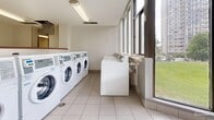 Located at 2 Silver Maple Crt in Brampton, Ontario, this shared laundry facility is a practical and well-organized amenity space spread across a single floor. The space is divided into two distinct areas connected by a tiled corridor with light-coloured flooring throughout.

The main laundry room is equipped with a row of front-loading washing machines alongside top-loading units and dryers, providing ample capacity for residents. Large windows line one wall, allowing natural light to fill the room and offering views of the surrounding green outdoor areas. Informational signage is posted throughout to guide users on proper machine operation and facility rules.

Adjacent to the laundry room is a dedicated folding and utility area, featuring a built-in counter with open shelving below — ideal for sorting and folding laundry. A wall-mounted display with a payment or information terminal is also present in this area. The space is accessed through a framed glass-panel door, lending a clean, semi-transparent partition between the corridor and the workspace.

A separate utility room houses electrical panels, a cork notice board for community postings, and a wall-mounted television. Fluorescent lighting runs across textured ceilings throughout the facility, ensuring bright, even illumination. The overall design is functional and clean, reflecting a well-kept shared amenity space suitable for everyday residential use.