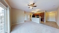 This single-floor residence features an open-concept layout that seamlessly connects the living area to the kitchen. The living space is finished with neutral-toned carpet and warm beige walls, creating a clean, understated backdrop. A ceiling fan with a built-in light fixture is centrally positioned overhead, providing both airflow and ambient lighting throughout the room. A doorway visible along one wall leads to additional spaces within the unit, while a utility panel is neatly recessed nearby.

The kitchen is defined by rich, dark wood cabinetry that extends to the upper reaches of the wall, offering generous storage. Black appliances, including a refrigerator and a microwave mounted above the range, complement the cabinetry's deep tones. A breakfast bar with a light-colored countertop serves as a natural divider between the kitchen and living area, providing a casual dining or workspace option. The kitchen is finished with tile flooring, offering a practical contrast to the carpeted living space. Overall, the layout is functional and cohesive, with consistent finishes that give the space a unified, move-in-ready feel.
