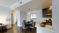This single-floor residence offers a thoughtfully designed open-concept layout that integrates the kitchen, dining, and living areas into a cohesive and functional space. The kitchen is outfitted with dark espresso cabinetry, light stone countertops, and a peninsula that doubles as a prep and serving area — providing both style and practicality. Adjacent to the kitchen, the dining area accommodates a round dark-wood table with upholstered grey chairs, lit by a warm pendant light that adds an inviting ambiance. A pass-through window connects the dining space to the bedroom beyond, enhancing the sense of openness and allowing natural light to flow throughout. Rich dark hardwood flooring runs continuously through the main living areas, offering a warm contrast to the crisp white walls and crown molding details. The bedroom, visible through the pass-through, is well-lit and neatly appointed. A bathroom is accessible from the hallway, featuring classic white fixtures and clean finishes. The overall design balances modern aesthetics with practical living, creating a space that feels both comfortable and refined.
