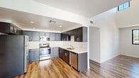 This two-story space showcases a contemporary design throughout, with wood-look flooring, recessed lighting, and a clean, neutral color palette that creates a bright and cohesive atmosphere. The kitchen is a standout feature, equipped with stainless steel appliances including a range, microwave, refrigerator, and dishwasher. Two-tone cabinetry — combining gray shaker-style lower cabinets with darker upper cabinets — frames light quartz countertops, while an L-shaped layout provides ample workspace and storage. The kitchen opens toward the living and dining areas, which benefit from high ceilings and direct access to an outdoor space via glass sliding doors.

Bedrooms are generously proportioned, with large black-framed windows that invite natural light and provide views of surrounding greenery. The bathroom features a dual-sink vanity with white shaker cabinetry, quartz countertops, oversized mirrors, and wood-look tile flooring.

A notable highlight is the expansive private rooftop deck, finished with a smooth concrete surface and framed by white stucco walls and terracotta tile rooflines. The deck enjoys open sky views and access through glass doors, making it an ideal outdoor retreat.
