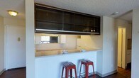 This single-story space offers a practical and open layout designed for comfortable everyday living. The kitchen is a standout feature, showcasing a peninsula-style breakfast bar topped with a light granite-look countertop, perfect for casual dining or entertaining. Two bold red metal stools add a pop of color and a modern industrial touch to the seating area. Above, dark-toned upper cabinets with glass-panel sliding doors provide ample storage while maintaining a sleek, contemporary aesthetic. The kitchen features a pass-through window that opens the space and promotes a sense of connectivity between rooms. White appliances and light-colored walls keep the kitchen feeling bright and clean, while under-cabinet lighting adds a warm, functional glow to the workspace. The textured ceiling adds a classic finish throughout the space. Dark hardwood-style flooring anchors the overall design, creating a pleasing contrast against the lighter tones of the countertops and walls. The overall ambiance is casual yet stylish, blending functional design elements with approachable, everyday comfort.