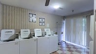 This single-floor shared laundry facility is designed with functionality and convenience in mind. The room features a row of top-load washing machines lined along one wall, each accompanied by informational and instructional notices posted above for user reference. The machines are well-spaced, allowing for comfortable access and ease of use.

The flooring consists of a classic checkered tile pattern in pink and white tones, adding a modest visual character to the utilitarian space. The walls are finished with a subtle striped wallpaper on the upper half, complemented by a lighter lower section, creating a clean and orderly aesthetic.

Overhead, a ceiling fan provides consistent air circulation throughout the room, supporting ventilation during use. A flush-mounted ceiling light ensures the space is well-lit. Natural light also enters through a large sliding glass door or window panel on one side of the room, brightening the interior and offering a connection to the outdoors.

A garment hanging rack positioned near the entry area adds a practical touch for air-drying or staging clothing. The overall layout is straightforward and efficient, making this laundry room a functional shared amenity suited for regular, everyday use.