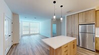 Located at 2311 Dulles Station Blvd in Herndon, Virginia, this single-floor residence showcases a clean, contemporary design with an open and airy layout. The kitchen serves as a centerpiece of the living space, featuring light wood cabinetry, white quartz countertops, a subway tile backsplash, stainless steel appliances, and a central island with pendant lighting overhead. Track lighting adds a modern touch, while the kitchen flows seamlessly into the open living area, which benefits from large windows that invite abundant natural light and wide-plank wood-style flooring underfoot. The bedrooms offer plush carpeting, generous wall space, and ample natural light from well-placed windows. One bedroom features double doors leading to an expansive walk-in closet with wire shelving systems, providing substantial storage capacity. The bathroom is finished with large-format neutral tiles, a soaking tub with a curved shower rod, a wood-accented vanity with a white quartz countertop, and an undermount sink. Throughout the residence, crisp white walls, clean lines, and consistent use of light wood tones create a cohesive, modern aesthetic that is both functional and inviting.