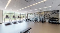 This fitness center offers a comprehensive workout environment spread across a single level, thoughtfully designed to accommodate a wide range of exercise needs. The main workout area is lined with floor-to-ceiling windows along one wall, flooding the space with natural light and providing views of surrounding greenery. The room features a drop ceiling with recessed linear lighting, smooth rubber flooring, and a striking full-wall graphic mural depicting vintage fitness and cycling imagery alongside the motivational phrase, "The Stronger You Are, The Better You Feel." Cardio equipment, including treadmills, elliptical trainers, and rowing machines, is arranged to face the expansive windows. Free weights, a cable machine, and adjustable benches are also available for strength training. A large wall-length mirror and vibrant yellow accent wall add visual energy to one section of the gym. Adjacent to the main floor is a dedicated studio space with hardwood flooring, glass partition walls, and an open layout ideal for stretching, yoga, or group exercise. Medicine balls and additional accessories are neatly stored along the walls. The overall design is clean, contemporary, and energizing.