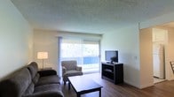 This single-floor space centers around a well-lit living area that draws immediate attention with its large sliding glass door, framed by vertical blinds that filter natural light while offering views of the surrounding greenery outside. The living area is designed for both comfort and functionality, featuring hardwood-style flooring that runs throughout and adds warmth to the space. A textured ceiling provides a classic finish overhead. The room comfortably accommodates a seating arrangement that includes a sofa, an upholstered armchair, and a rectangular coffee table, all oriented toward an entertainment unit housing a flat-screen television. The dark-toned media console provides a contrast against the neutral walls, contributing to the room's understated, practical aesthetic. A floor lamp in the corner supplements the natural light that pours in through the sliding door. The sliding door itself serves a dual purpose, functioning as both a source of daylight and a direct access point to the outdoor area visible beyond. Overall, the living space offers a functional and comfortable layout with a clean, straightforward design suited for everyday living.