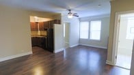 This single-floor space offers a thoughtfully designed open-concept layout that seamlessly connects the living area and kitchen, creating a cohesive and functional environment. Rich, dark hardwood flooring runs throughout, adding warmth and a polished aesthetic to the space.

The living area is bright and airy, benefiting from multiple windows that allow generous natural light to fill the room. A ceiling fan is centrally positioned overhead, providing both practical airflow and a subtle design element. Clean white walls and crisp white baseboards complement the flooring, lending the space a fresh, well-maintained appearance.

The kitchen is defined by dark-stained wood cabinetry that extends to the upper and lower walls, offering ample storage. Dark stone countertops provide a durable and visually striking workspace, while black appliances — including a refrigerator and gas range — lend a uniform, modern finish to the cooking area. The kitchen flows naturally into the main living space, making it ideal for entertaining or everyday use.

A doorway visible from the main area leads to additional rooms, suggesting a practical and connected floor plan. Overall, the space presents a cohesive blend of dark, rich finishes against light-toned walls, creating an inviting and well-balanced interior.