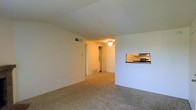 This single-floor space features an open living area finished with wall-to-wall carpet in a neutral beige tone, creating a clean and understated foundation throughout. The living room is generously proportioned, with smooth white walls and a textured ceiling that add a simple, classic character to the space. A ceiling fan is mounted overhead in the adjoining area, providing both ventilation and a subtle focal point. Natural airflow is supported by a visible floor vent along the baseboard. A defining architectural feature is the pass-through window cut into the wall between the living area and the kitchen, allowing visual connectivity between the two spaces without fully opening the floor plan. The kitchen, glimpsed through this opening, includes cabinetry in a light finish and a dark countertop, suggesting a compact but functional cooking space. A doorway visible in the background leads further into the unit, hinting at additional rooms beyond. The overall ambiance is neutral and move-in ready, with a practical layout that emphasizes efficient use of space and ease of flow between the living and kitchen areas.