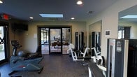 Located in Saddle River, this single-floor fitness facility is thoughtfully designed to support an active lifestyle in a clean, modern environment. The workout area is equipped with a selection of strength and cardio machines, including resistance training equipment, a rowing machine, and a weight bench, all arranged on durable rubberized flooring ideal for high-performance exercise. A large wall-mounted mirror enhances the sense of space and allows for proper form checking during workouts. Natural light filters in through a centrally placed skylight, complementing the recessed ceiling lighting to create a bright and energizing atmosphere. The dark-toned ceiling adds a sleek, contemporary contrast to the neutral walls. A set of glass-paneled double doors connects the fitness area to an adjacent lounge or meeting space, which is visible through the doors and features warm seating, a decorative area rug, and views of lush greenery beyond. This seamless connection between the active and social spaces makes the facility well-suited for both individual training and community-oriented use. The overall design balances functionality with a refined aesthetic, offering a professional-grade workout environment.
