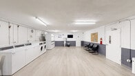 This community laundry facility is a spacious, single-floor space designed to efficiently serve its users. The interior features light wood-style flooring and white-painted concrete block walls accented with a gray wainscoting band, giving the room a clean, functional aesthetic. Bright fluorescent lighting runs across the ceiling, ensuring the entire space is well-lit throughout the day.

Along one side of the room, a comprehensive bank of commercial-grade top-load and front-load washing machines, as well as stacked dryer units, lines the wall. A utility sink area provides additional convenience for hand-washing or pre-treating garments. Exposed overhead plumbing is neatly routed along the walls, reflecting the utilitarian nature of the space.

On the opposite side, a dedicated waiting area features several black cushioned chairs arranged along the wall, alongside a wall-mounted flat-screen television for resident entertainment. A cork community bulletin board labeled "WLA Community Board" is prominently displayed, serving as an informational hub for residents. A vending machine and a recycling station are also available, adding further convenience.

The open floor plan allows for easy movement throughout the space, while a clearly marked exit door ensures safe and accessible egress. Overall, the facility is thoughtfully organized to provide a practical and comfortable laundry experience.